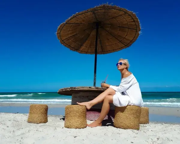 Kobieta w białej sukience relaksuje się pod parasolem na plaży, popijając napój. Idealne tunezja wakacje.