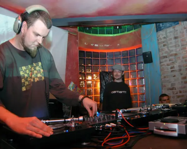 DJ Maceo Wyro w akcji, miksuje muzykę w klubie. W tle widać innych DJ-ów i kolorowe oświetlenie.