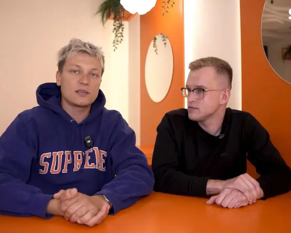 Dwóch mężczyzn siedzi przy pomarańczowym stole. Jeden w niebieskiej bluzie z napisem "SUPREME", drugi w czarnej koszulce. Rozmawiają o kebabie friza.