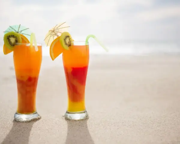 Dwa drinki Malibu z sokiem pomarańczowym, ozdobione plasterkami kiwi i pomarańczy, stoją na piaszczystej plaży.