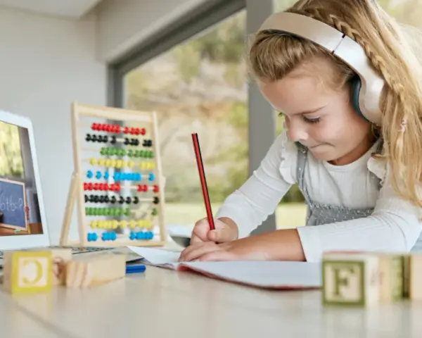 Dziewczynka w słuchawkach uczy się matematyki online, pisząc w zeszycie. Na ekranie laptopa nauczycielka pokazuje zadanie.