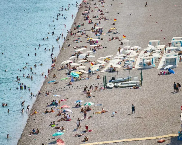 Tłum ludzi odpoczywa na kamienistej plaży w Finike. Niektórzy kąpią się w morzu, inni opalają się pod parasolami.