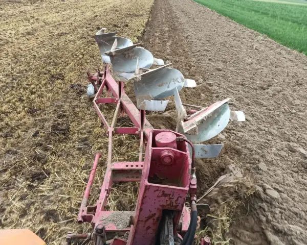 Czerwony pług z kilkoma lemieszami pracuje na polu, odwracając ziemię. Trwa regulacja pługa, przygotowując pole do siewu.
