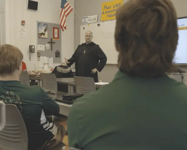 A monastery leader in black robes addresses students in a classroom.