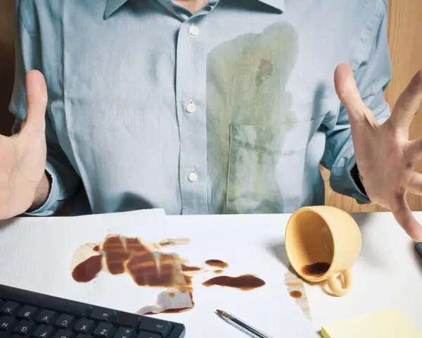 Mann mit verschüttetem Kaffee auf Hemd und Schreibtisch. Eingetrocknete Kaffeeflecken entfernen ist eine Herausforderung.