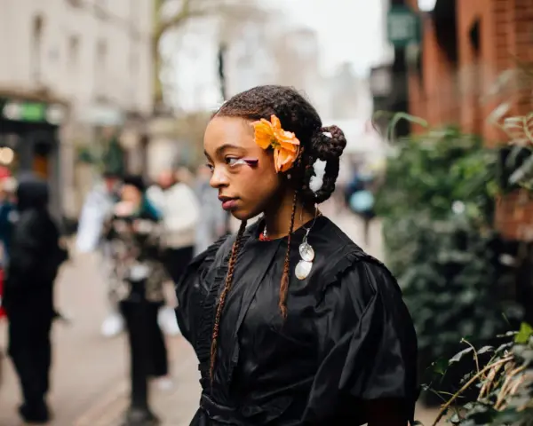 Ragazza con trecce davanti e capelli sciolti, ornata da un fiore arancione e makeup audace. Indossa un abito nero con dettagli voluminosi.