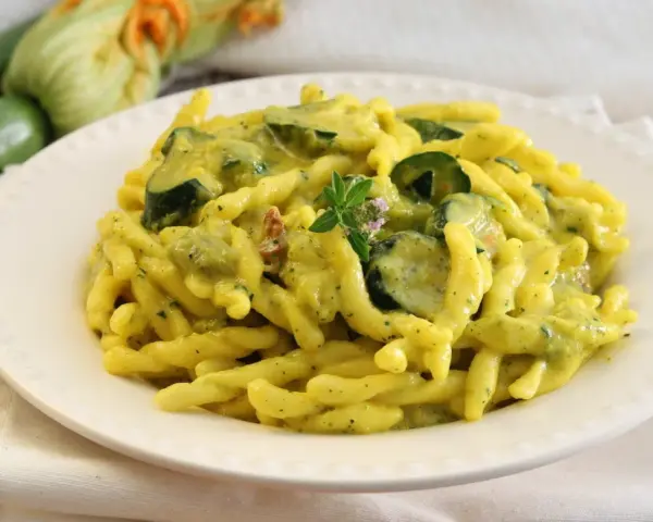 Piatto di pasta con zucchine e zafferano, guarnito con un rametto di erbe aromatiche. Sullo sfondo zucchine fresche e fiori di zucca.