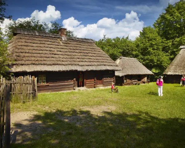 Skansen Chorzów bilety: drewniane chaty kryte strzechą, dzieci bawią się na trawie, zwiedzający podziwiają architekturę.