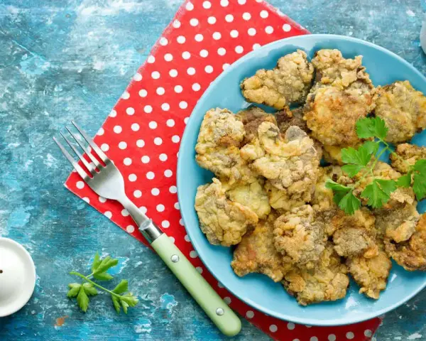 Piatto di fegatini di pollo impanati e fritti, guarniti con prezzemolo. Accanto, una forchetta e un sale e pepe.