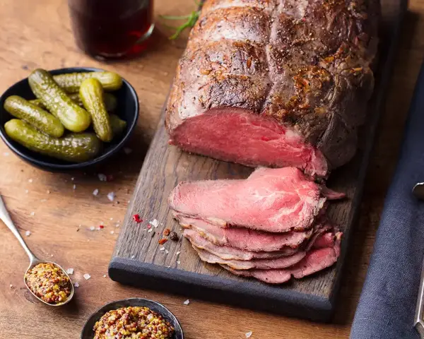 Un délicieux roti de boeuf, tranché et servi avec des cornichons, de la moutarde et une boisson foncée.