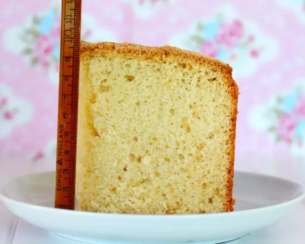 Rebanada de bizcocho casero esponjoso, con una regla de madera al lado para medir su altura. Fondo rosa con flores.