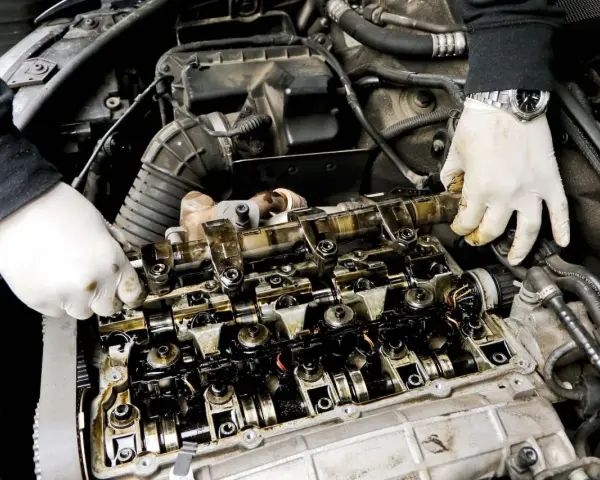 Mechanik w białych rękawiczkach sprawdza wałek rozrządu, szukając objawów zużycia. Widoczne są elementy silnika i przewody.