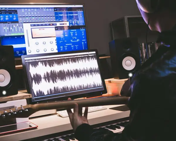 Tworzenie muzyki w domowym studiu. Na ekranach widać programy do edycji dźwięku, a w rękach gitarzysty gryf gitary.