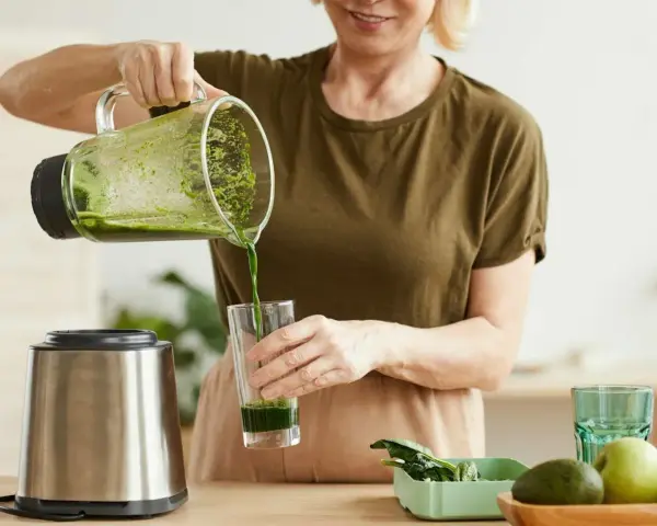 Kobieta nalewa zielony koktajl położnej do szklanki. Na blacie widać blender, awokado i jabłko.