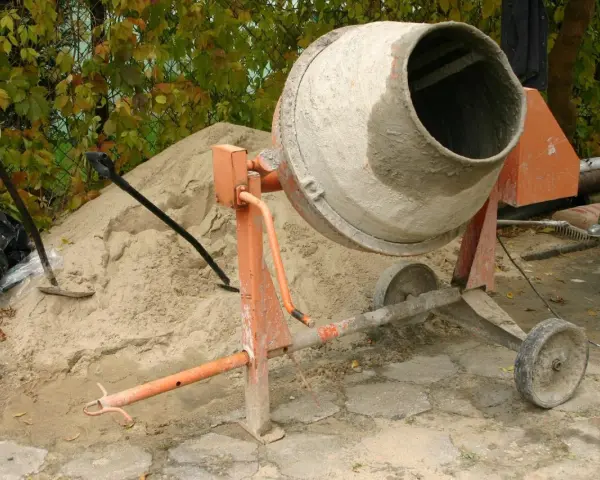 Betoniarka 200l czeka na piasek i cement. Ile cementu na betoniarkę 200l? Obok łopata i kapelusz.