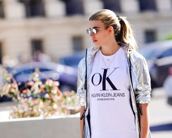 Kobieta w srebrnej kurtce CK damskiej i białej koszulce z logo Calvin Klein Jeans.