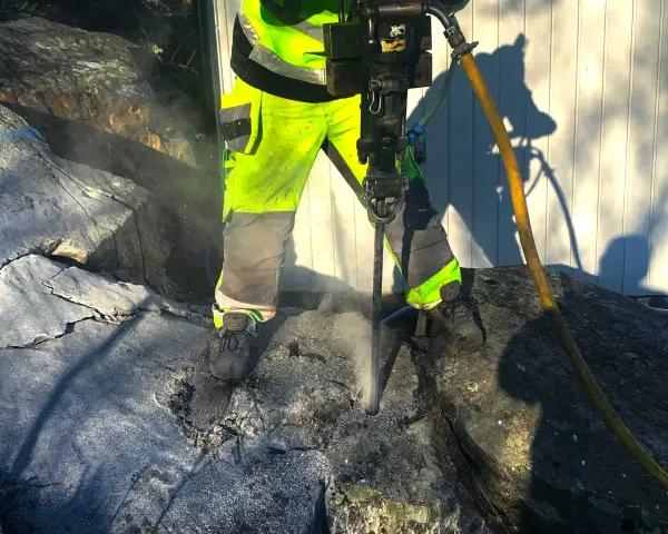 Arbetare med reflexkläder borrar i berg med en kraftig borrhammare. Dammet yr runt borren.