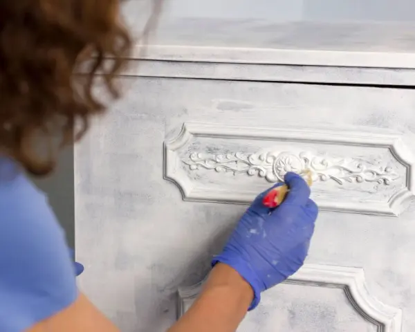 Mano enguantada aplica pintura a muebles pintados a la tiza en dos colores, dando un acabado desgastado y elegante.