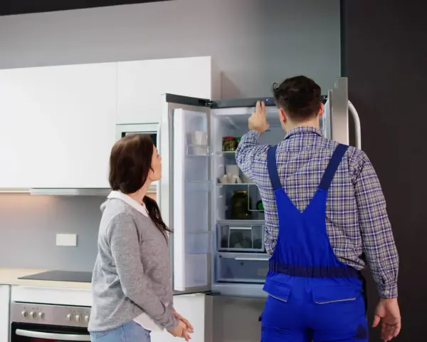 Un technicien en salopette bleue vérifie un frigo qui claque, tandis qu'une femme observe attentivement.