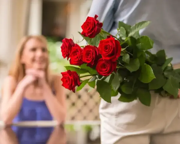 Mężczyzna z bukietem czerwonych róż za plecami, zbliża się do uśmiechniętej kobiety. To piękny gest, jak na 60 lat po ślubie, jakie to gody!