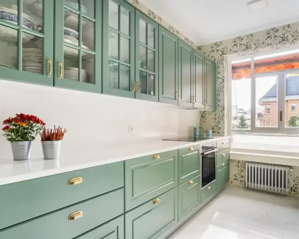 Encantadoras cocinas verdes vintage con armarios de cristal y detalles dorados. Flores rojas y naranjas adornan la encimera blanca.
