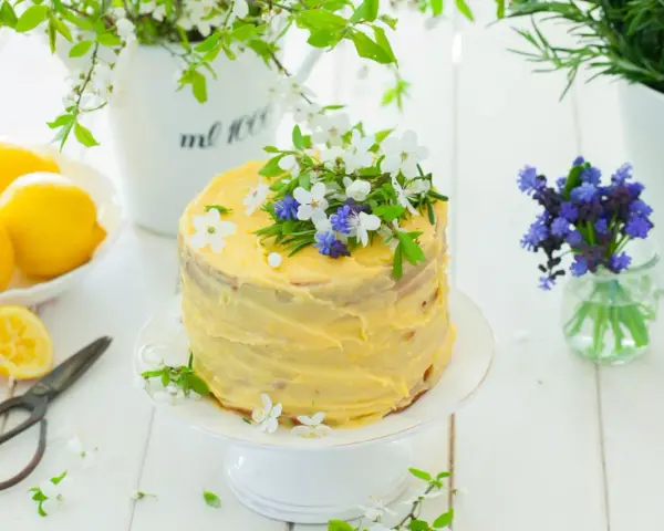Słoneczny tort cytrynowy z kremem lemon curd, ozdobiony świeżymi kwiatami i ziołami. Obok leżą cytryny i stare nożyczki.