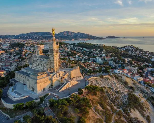 La basilique Notre-Dame de la Garde veille sur Marseille, baignée par la lumière dorée du soleil couchant. La mer et les collines environnantes s'étendent à perte de vue.