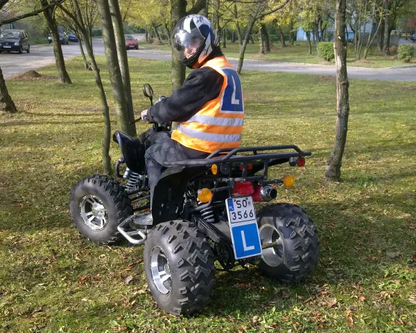 Młody kierowca w kamizelce odblaskowej i kasku uczy się jeździć quadem. Tablica z literą "L" wskazuje, że to kategoria B1.