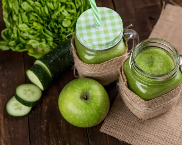 Dwa słoiki z zielonym smoothie, obok jabłko, plasterki ogórka i sałata. Orzeźwiający drink z ogórkiem na drewnianym tle.