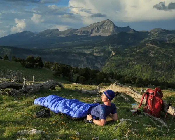 Mann liegt in blauer Biwakschachtel im Freien, umgeben von Berglandschaft. Rucksack und Wanderstöcke daneben.