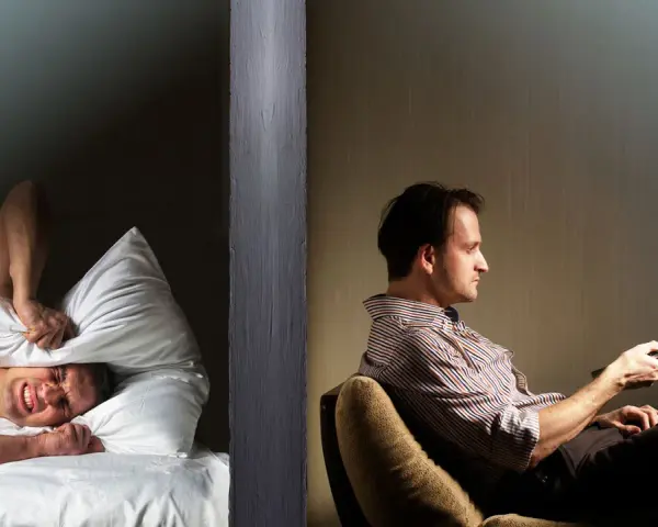 Hombre en cama tapa oídos con almohada, sufriendo por vecinos ruidosos. Al otro lado, otro hombre ve TV.