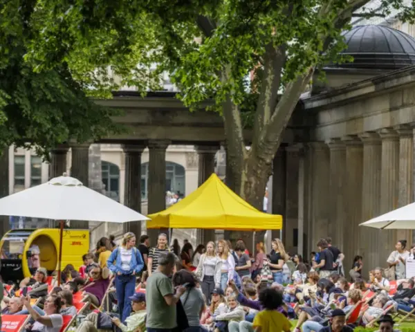 Menschen genießen entspannte **Veranstaltungen am Wochenende** im Freien vor einer Säulenfassade. Sonnenschirme und Zelte spenden Schatten.