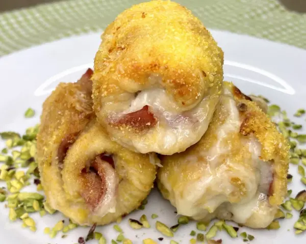 Tre involtini di pollo dorati e ripieni di formaggio filante e prosciutto, adagiati su un letto di pistacchi tritati.