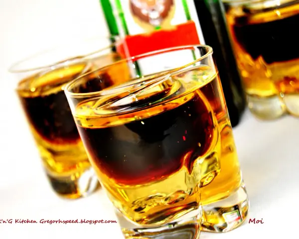 Dwa kieliszki z warstwowym drinkiem Jägermeister i Red Bull. Butelka Jägermeister w tle.