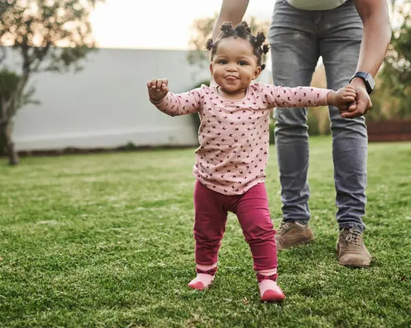 Ein kleines Mädchen mit lustigen Zöpfen macht seine ersten Schritte im Gras, gehalten von einer erwachsenen Hand. Wann laufen Babys? Heute ist ein guter Tag dafür!