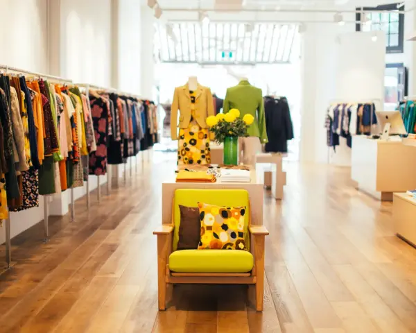 Interior de un showroom de ropa con percheros llenos de prendas coloridas y un sillón verde con cojines estampados.