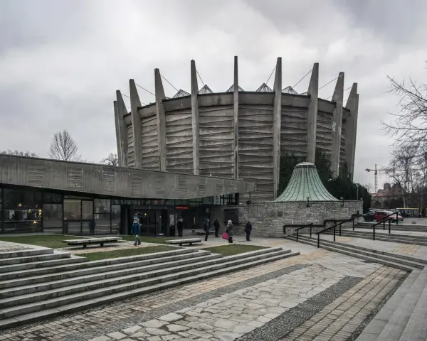 Monumentalny budynek w stylu brutalizm z betonowymi żebrami i szklaną fasadą. Schody prowadzą do wejścia, a wokół zielone trawniki.