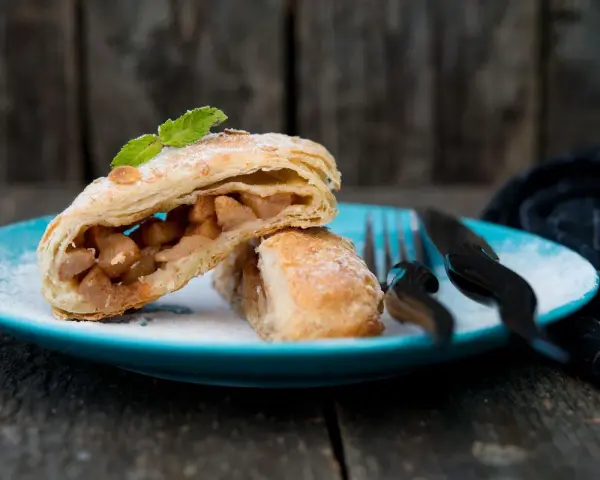 Delizioso strudel pere cioccolato e amaretti, con ripieno di mele e un tocco di menta, su un piatto blu cosparso di zucchero a velo.