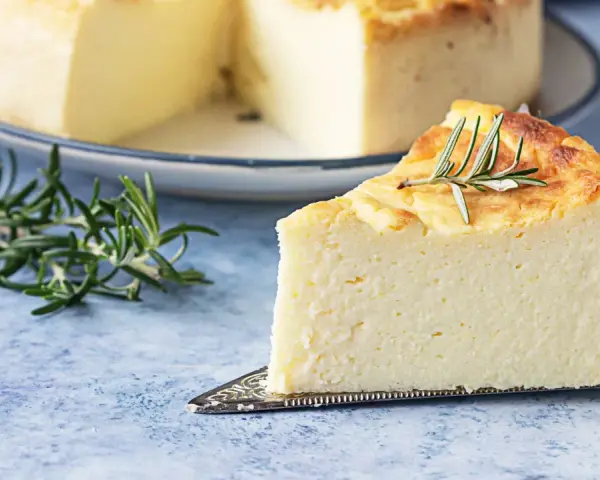 Una fetta di torta di ricotta soffice e dorata, guarnita con rosmarino, su un piatto da torta.