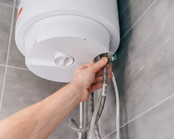 Mano che controlla la valvola scaldabagno perde. Tubi flessibili collegati a un boiler bianco su sfondo piastrellato.