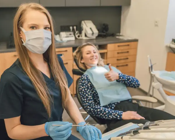 Uśmiechnięta pacjentka daje kciuk w górę podczas pierwszej wizyty u dentysty po latach. Dentystka w masce i rękawiczkach przygotowuje narzędzia.