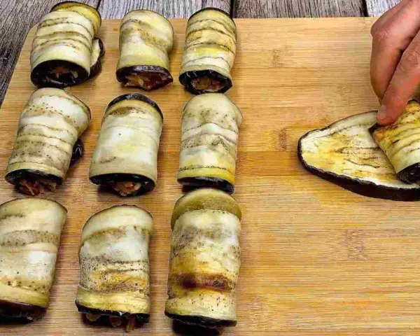Nove involtini di melanzane pronti, uno viene arrotolato su una fetta di melanzana grigliata.