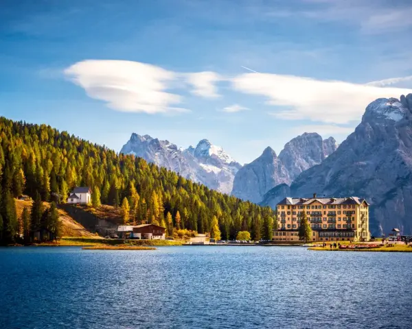 Laghi del Veneto in moto: un hotel giallo e una chiesetta bianca su un pendio boscoso, con montagne innevate sullo sfondo.