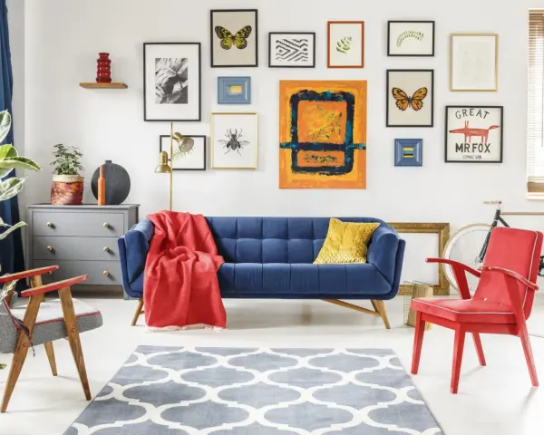 Salon w stylu eklektycznym: granatowa kanapa, czerwone fotele, galeria obrazów z motywami natury i abstrakcji, szara komoda, dywan z geometrycznym wzorem.