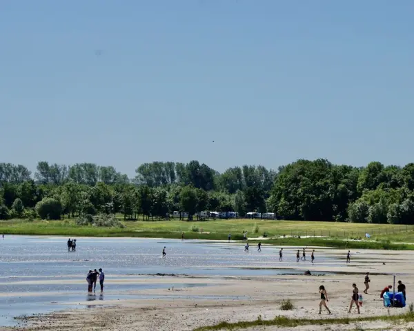 Menschen genießen einen sonnigen Tag am Strandbad Eldena. Einige baden im flachen Wasser, andere spazieren am Ufer entlang.