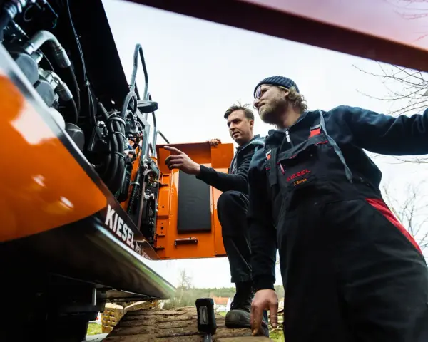 Dwóch mechaników w kombinezonach przeprowadza konserwację maszyn budowlanych. Jeden z nich wskazuje na silnik.