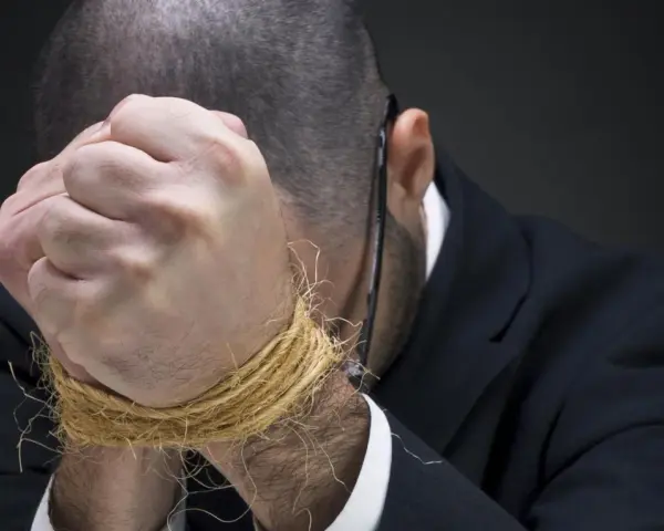 Mężczyzna w garniturze z rękami związanymi sznurkiem, pochylony.