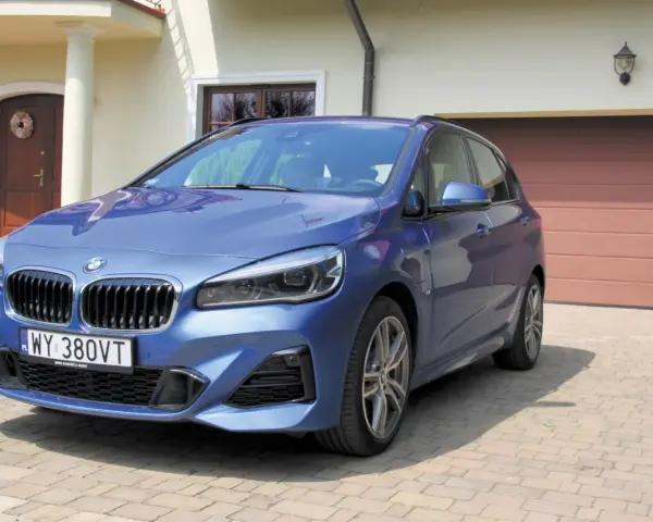 Niebieskie BMW 218i zaparkowane przed domem z brązową bramą garażową.