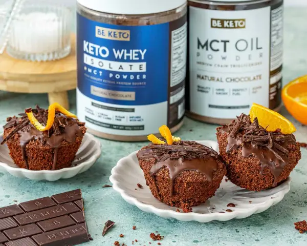 Pyszne keto muffiny z mąki migdałowej, polane czekoladą i ozdobione skórką pomarańczową. Obok stoją puszki z odżywką białkową i olejem MCT.