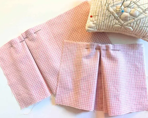 Tissu rose à carreaux avec des plis épinglés, montrant comment faire des plis en couture avec une fourchette. Coussin à épingles en arrière-plan.